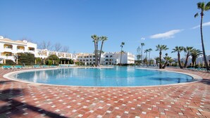 A swimming pool with a brick deck, surrounded by palm trees and a row of white buildings with balconies. - 'Golden' Apartment with Shared Pool, Wi-Fi and Air Conditioning (Cabanas de Tavira)