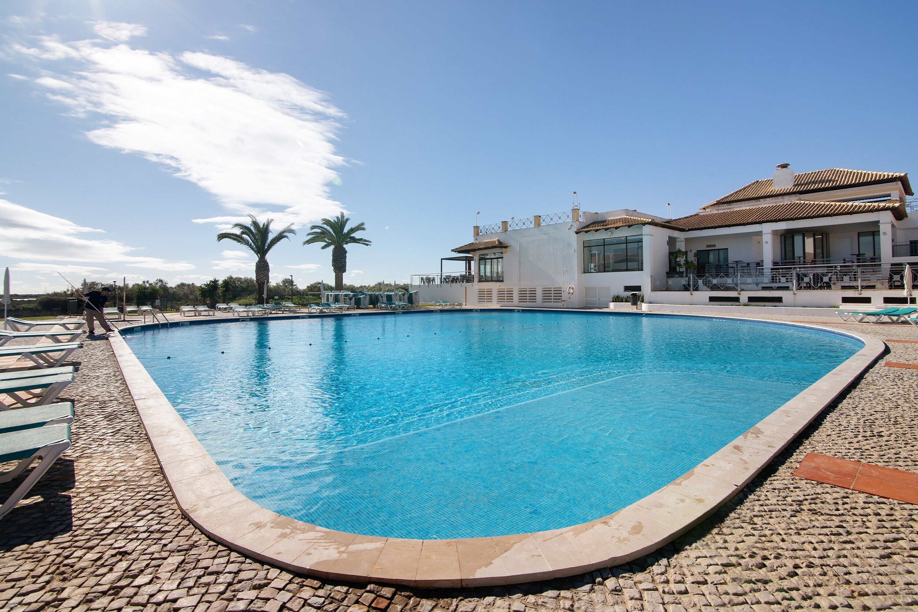 Une piscine avec des chaises longues, un bâtiment doté d’un toit en tuiles et des palmiers en arrière-plan.