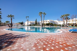 A swimming pool with a tiled deck, palm trees, and a row of white buildings with balconies.