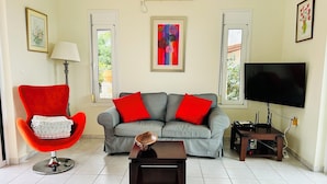A living room with a grey sofa, red cushions, a red armchair, a coffee table, a TV, and a framed painting on the wall. - Charming and serene villa with pool, lush garden, sea and mountain view (Vamos)