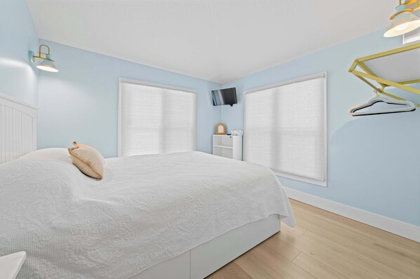 A bedroom with a bed, two windows with blinds, a TV mounted on the wall, and a hanging light fixture.