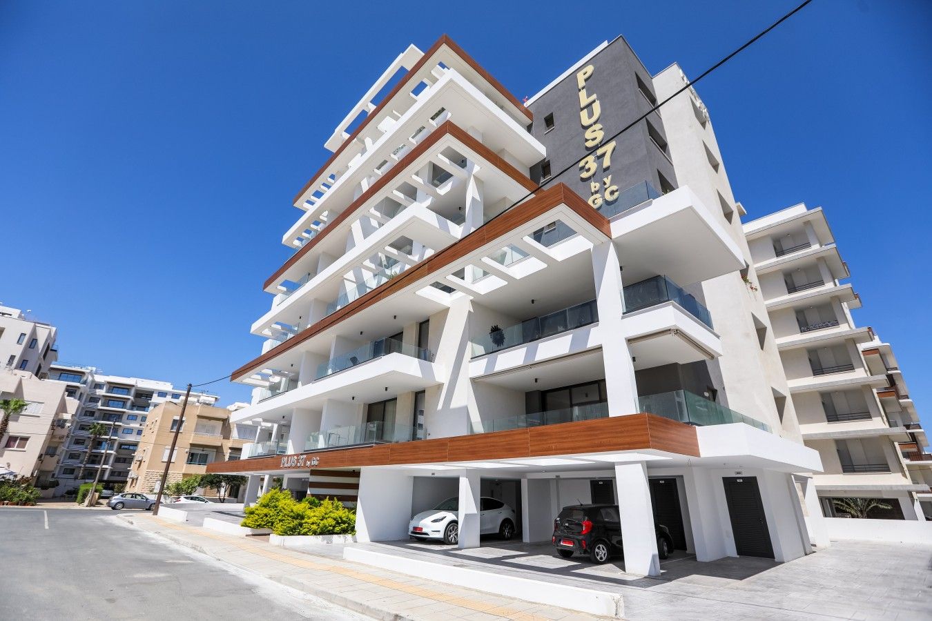 A multi-story hotel with a modern design, featuring balconies on each floor and a parking area in front.
