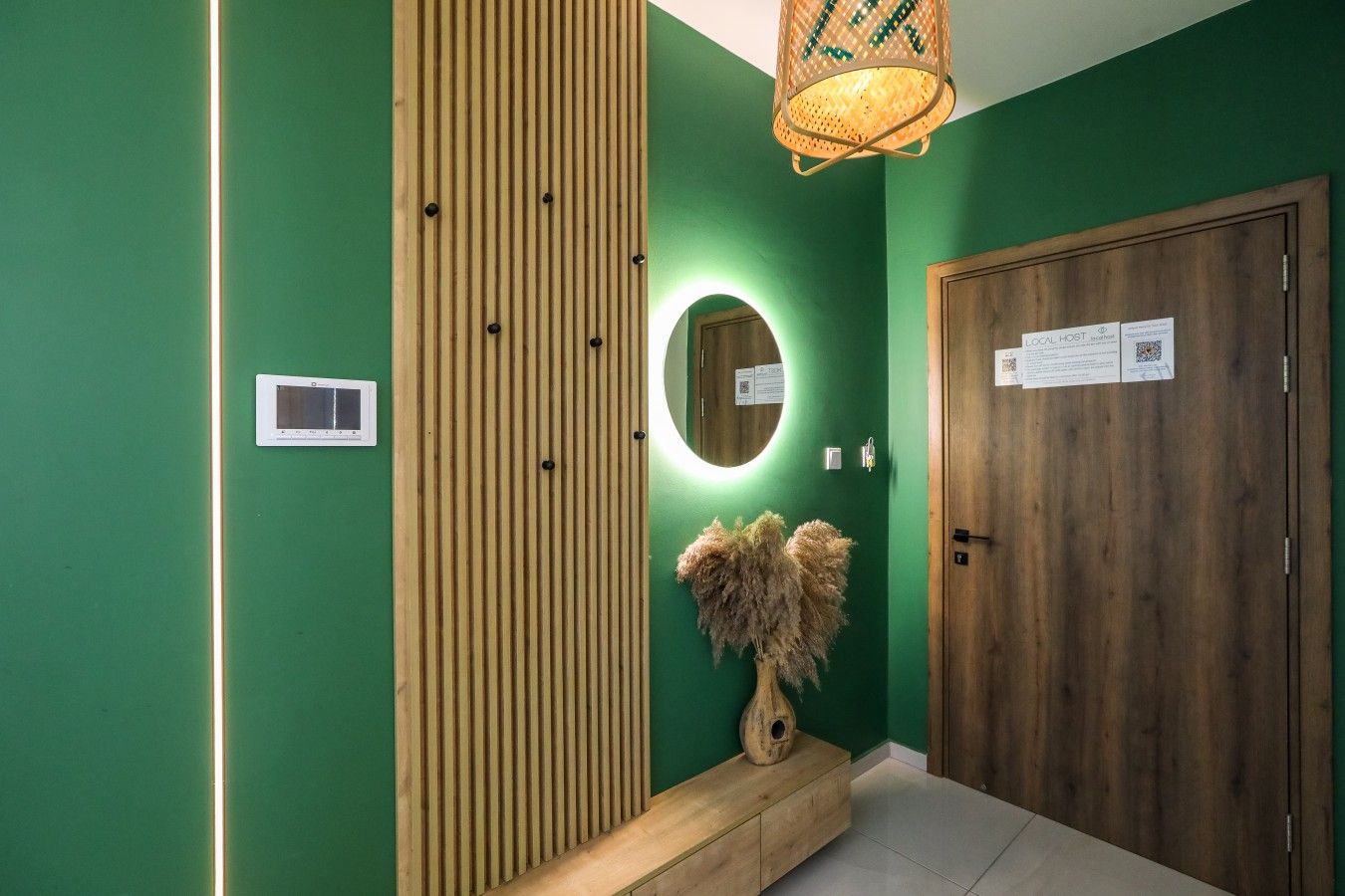 A hallway with a green wall, a wooden panel, a mirror, a vase with dried plants, and a door with a sign.