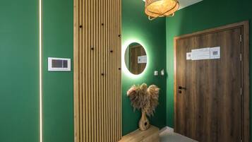 A hallway with a green wall, a wooden panel, a mirror, a vase with dried plants, and a door with a sign.