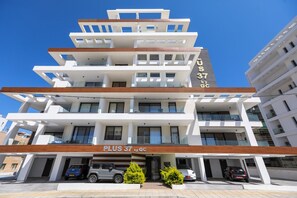 Un hôtel à plusieurs étages au design moderne, doté de balcons à chaque niveau, d’un parking où sont stationnées plusieurs voitures, et avec un ciel bleu azur en arrière-plan.
