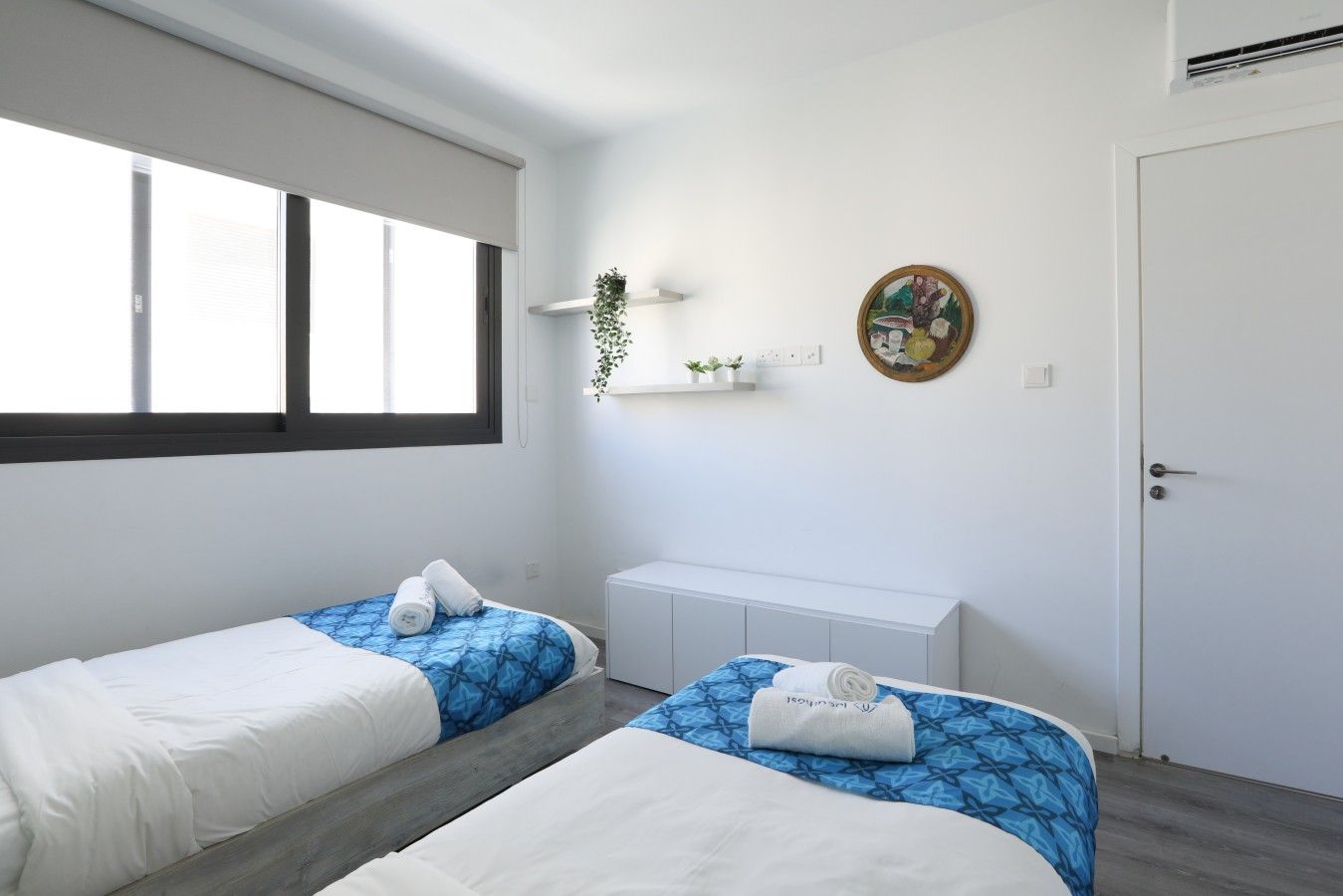 Une pièce avec deux lits, une fenêtre, une porte, une petite plante et une étagère fixée au mur.