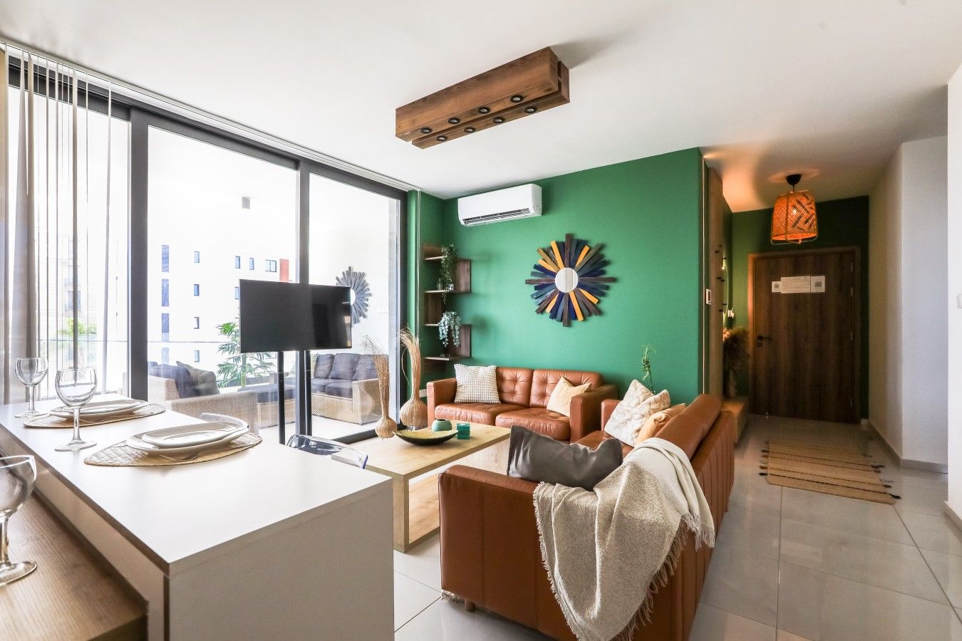 A modern living room with a large window, a flat-screen TV, a brown leather sofa, and a green accent wall.