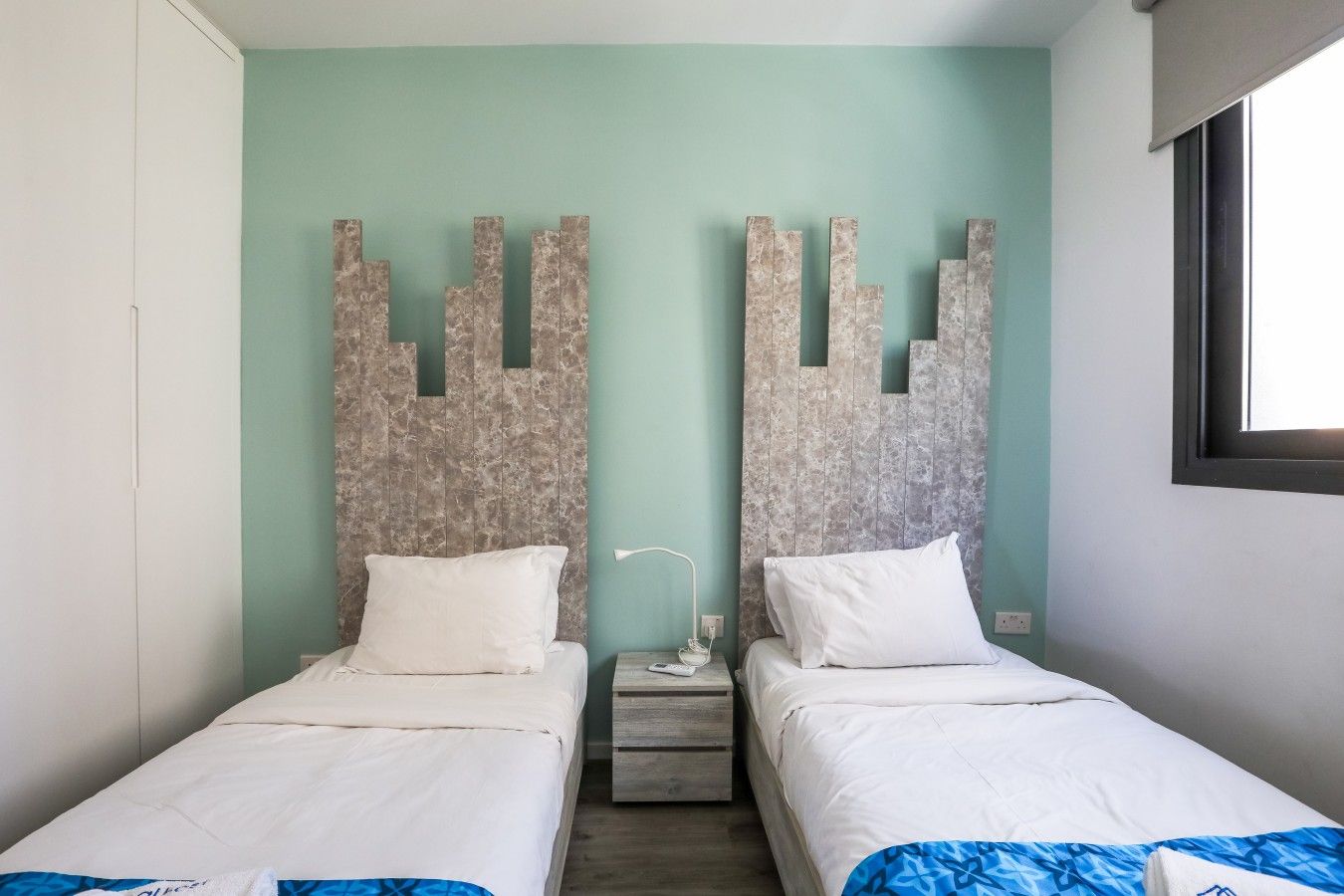 A room with two beds, a nightstand, and a wall-mounted light fixture.