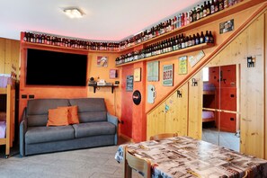 A cozy room with a sofa, a table with a newspaper-covered tablecloth, a TV, and a wall with shelves of bottles. - Apartment 'Taverna Relax Sara' with Mountain View, Private Terrace and Wi-Fi (Triangia)
