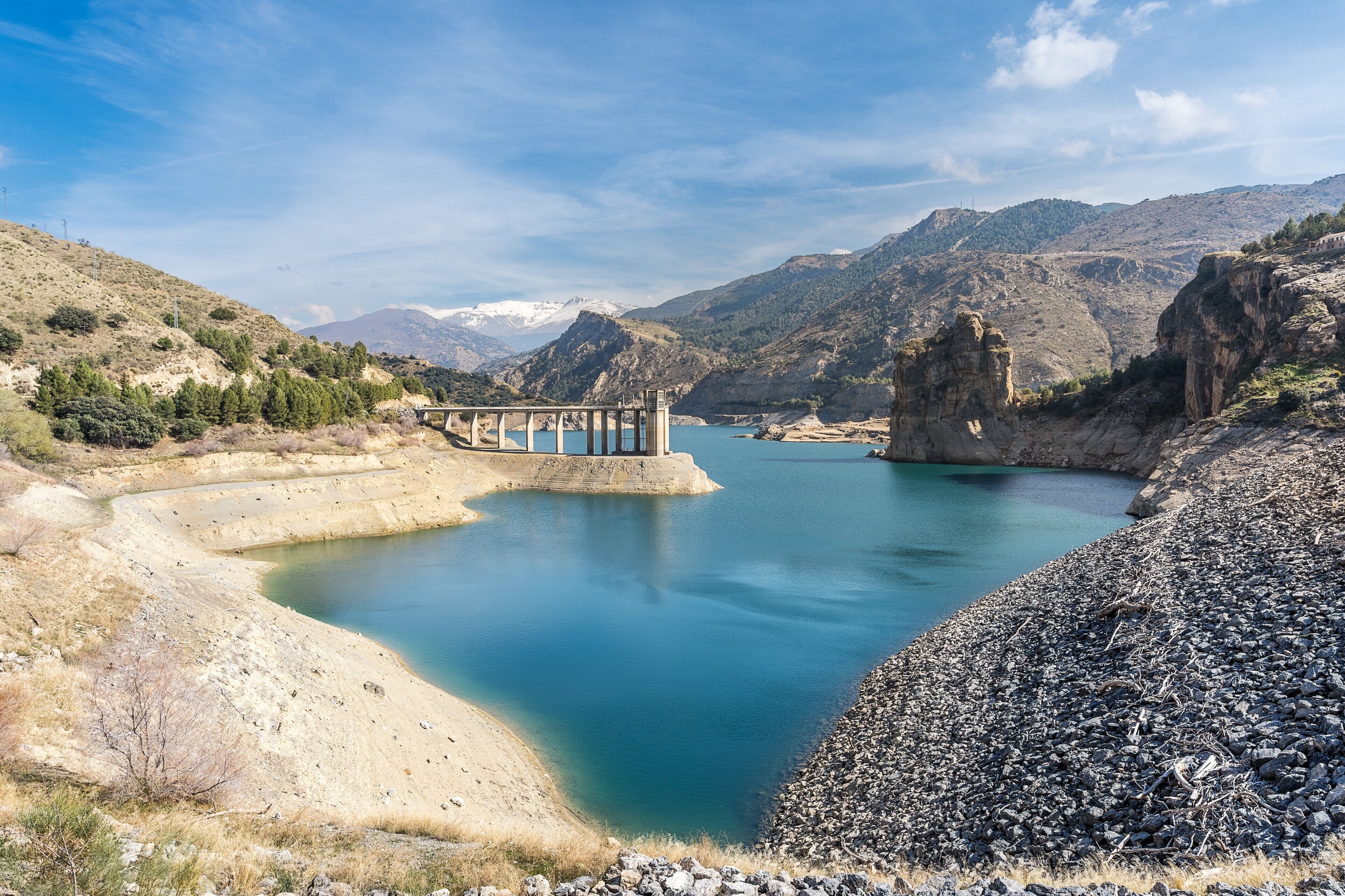 Una presa con un puente, agua tranquila, terreno rocoso y montañas al fondo.
