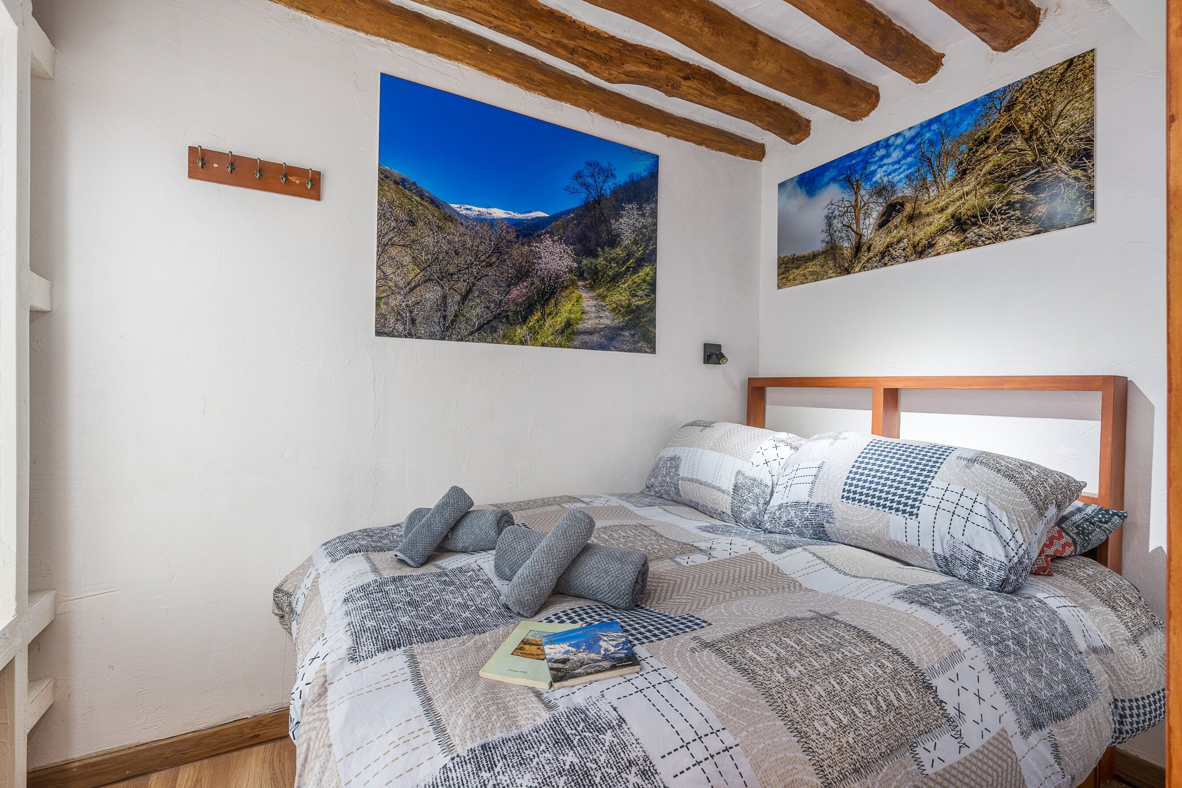 A bedroom with a bed, a wooden headboard, two wall-mounted paintings, and a small shelf.
