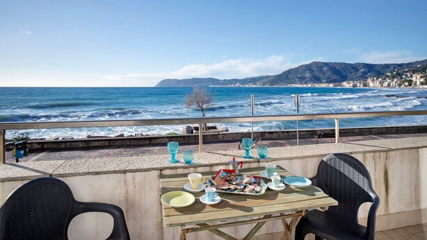 Een gedekte tafel met borden, bekers en een dienblad vol eten, met uitzicht op een strand met golven en een verre skyline.