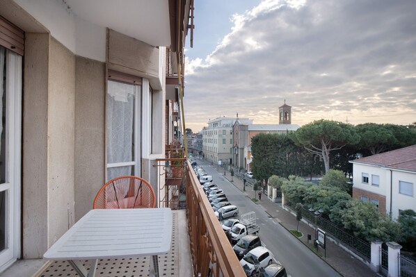 A balcony with a white table and orange chair, overlooking a street with parked cars and buildings.