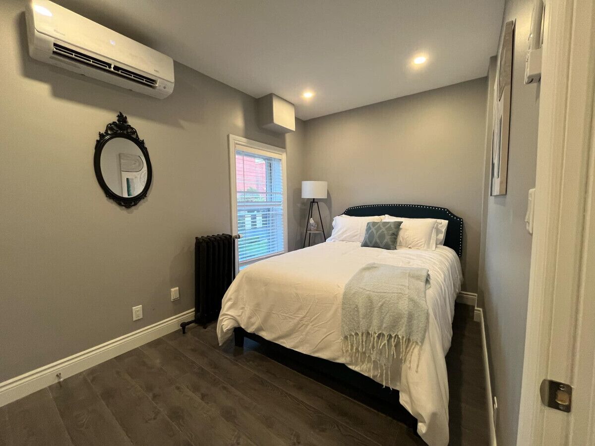 A bedroom with a bed, a mirror, an air conditioner, a radiator, and a window with blinds.