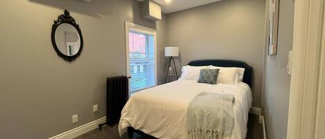 A bedroom with a bed, a mirror, an air conditioner, a radiator, and a window with blinds.