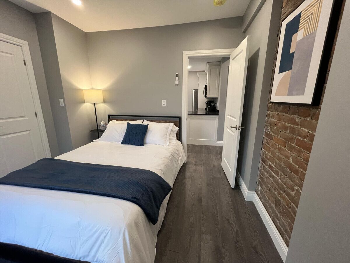 A bedroom with a bed, bedside table, lamp, and a view into the kitchen.