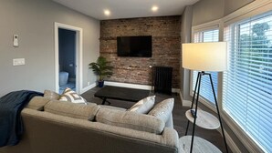 Ein modernes Wohnzimmer mit einer Couch, einem Couchtisch, einem an einer Ziegelwand montierten Fernseher, einer Stehlampe und einem Fenster mit Jalousien.