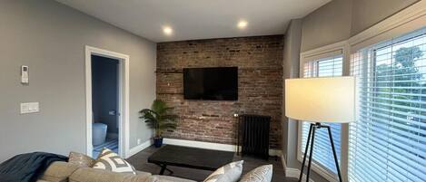 A modern living room with a sofa, a coffee table, a TV mounted on a brick wall, a floor lamp, and a window with blinds.