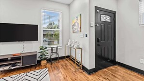 A living room with a flat-screen TV, a potted plant, a small stool, and a door with a decorative glass panel.