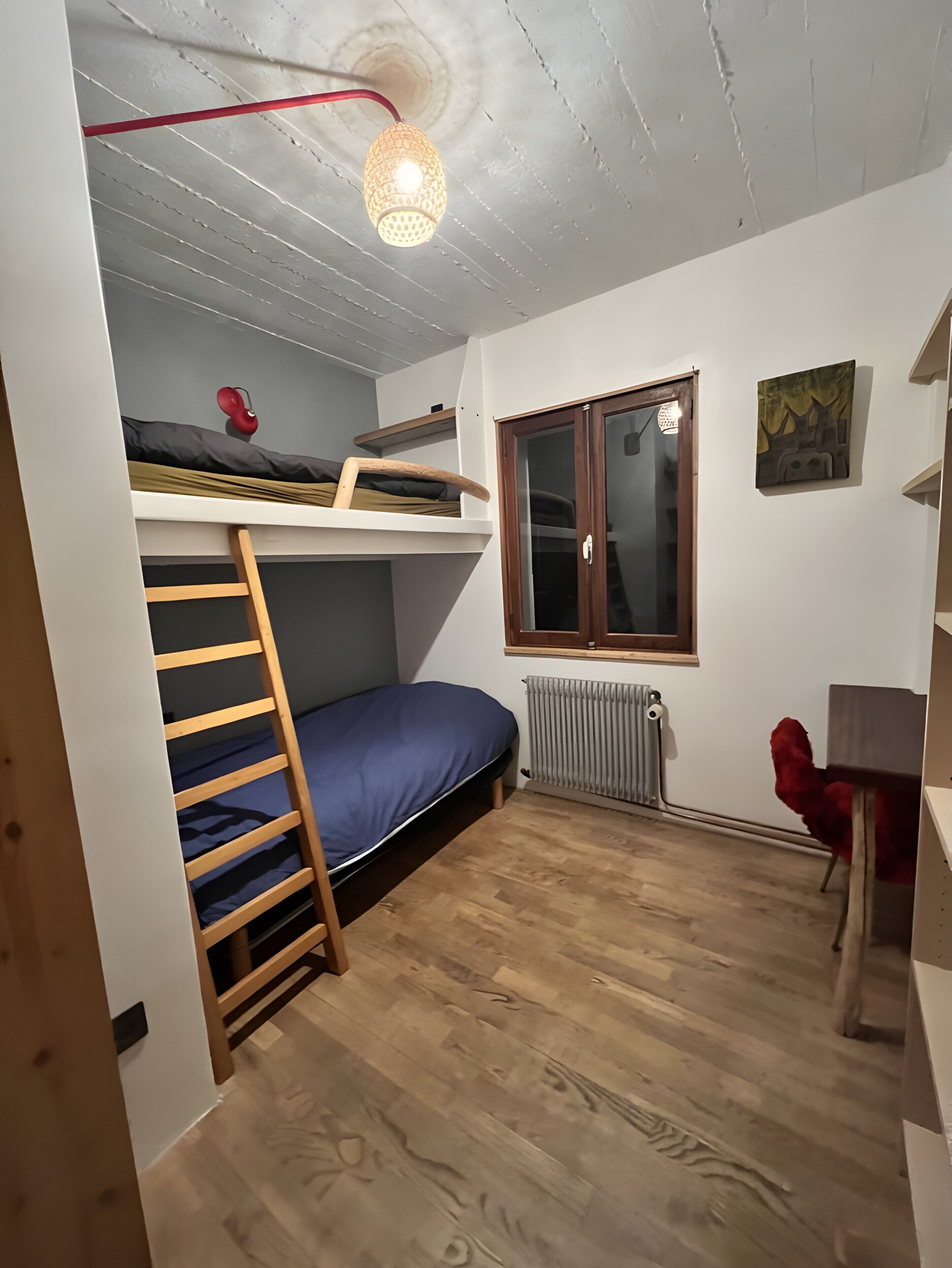 Une petite pièce comprenant un lit superposé, une échelle, un bureau, un radiateur et une fenêtre.