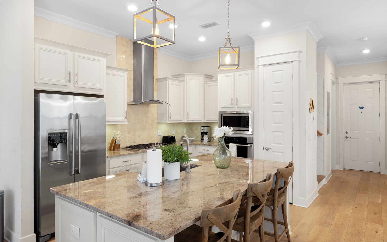 A modern kitchen with white cabinetry, stainless steel appliances, a marble countertop, and wooden chairs.