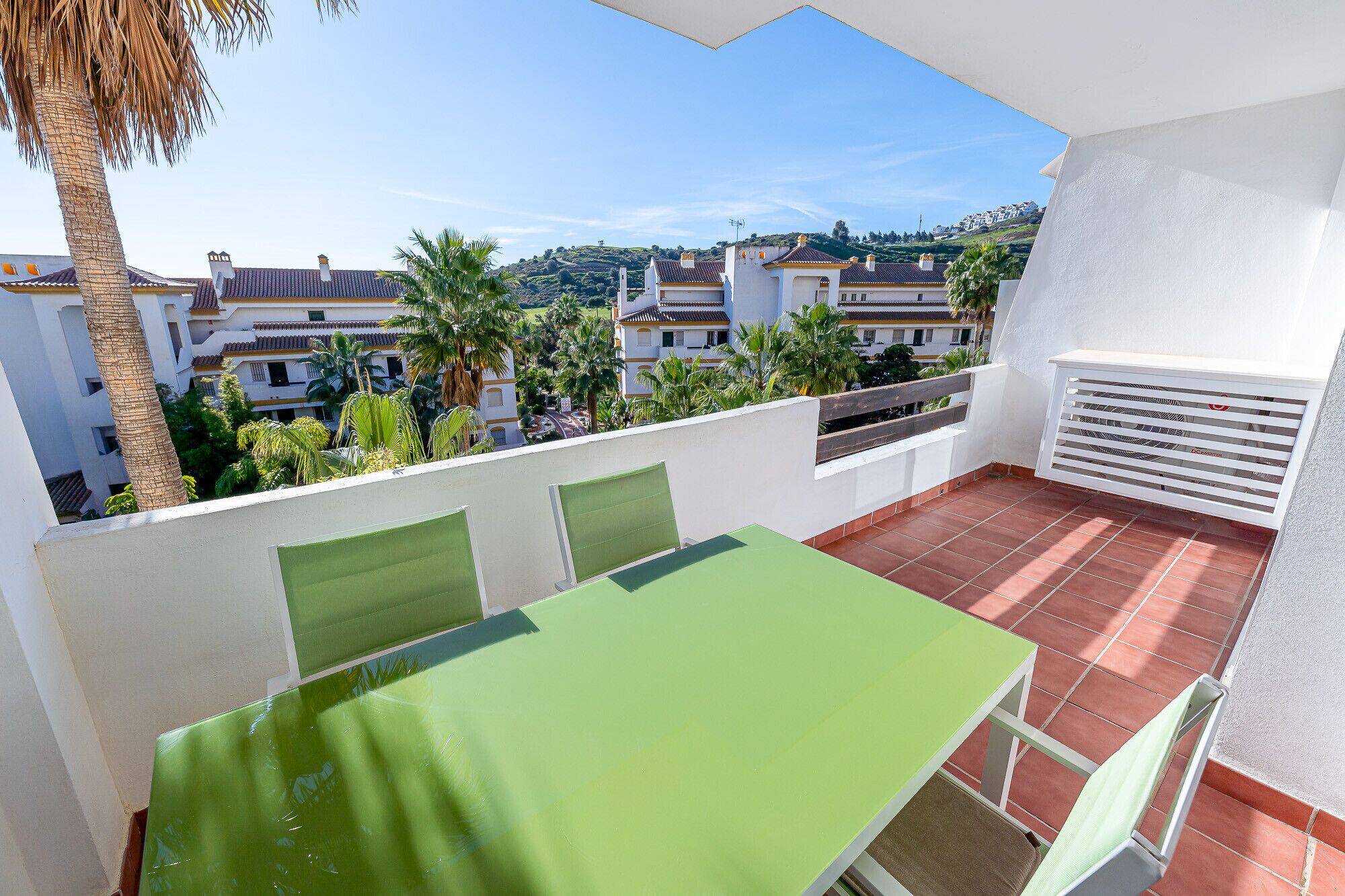 A balcony with a green table and chairs, a white wall, and a view of buildings and palm trees.