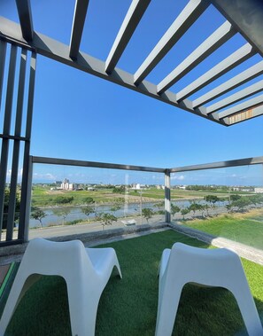Habitación con vista a un río y edificios, con dos sillas de plástico blancas y piso de césped artificial verde.