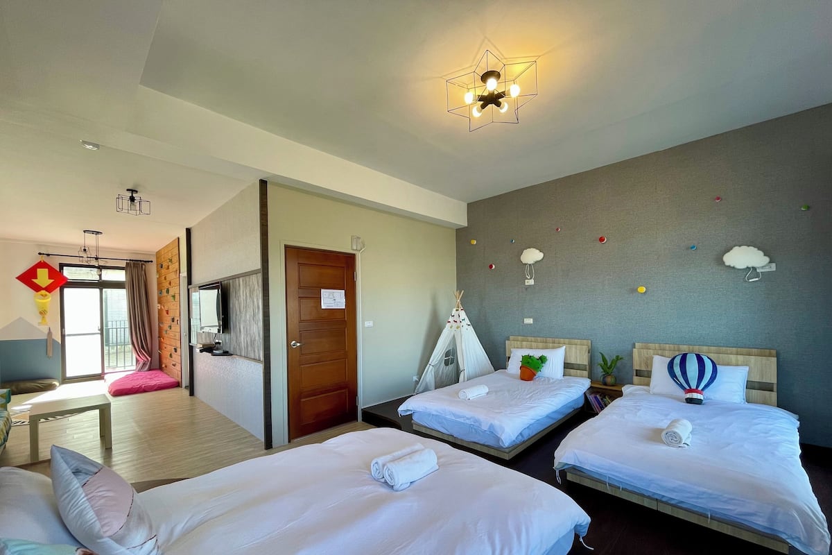 A hotel room with two beds, a wooden door, a window with curtains, a ceiling light fixture, and wall decorations including clouds and a hot air balloon.