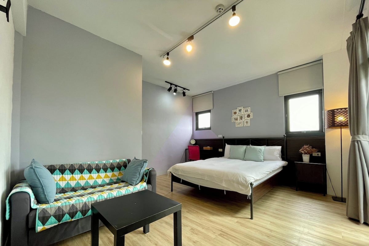 A modern, well-lit room with a bed, a sofa, a small table, and a window with curtains.
