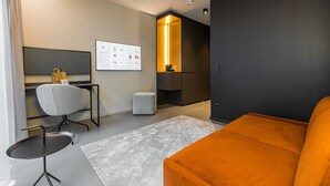Een moderne hotelkamer met een grijze bank, een klein rond tafeltje, een grijze fauteuil, een groot ingelijst schilderij aan de muur en een ingebouwde televisie.