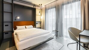 Een moderne hotelkamer met een bed, een bureau, een stoel en een groot raam met gordijnen.