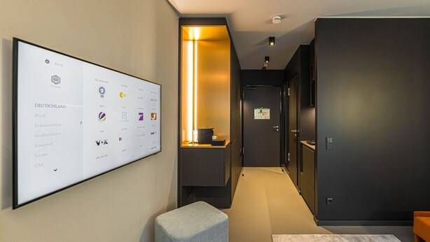 Een moderne hotelgang met een digitaal scherm waarop televisiezenders staan vermeld, een heldere gangverlichting en een deur met een bordje.