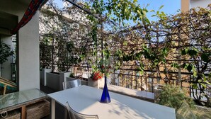 A balcony with a white table, a blue vase with flowers, and a view of a garden with a metal fence and trees. - Italianway - Venini 16 (Milan)