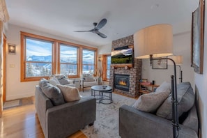 A living room with a fireplace, large windows, and a ceiling fan.