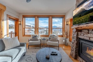 Ein gemütliches Wohnzimmer mit Kamin, großen Fenstern, einer Couch, zwei Sesseln und einem Couchtisch.