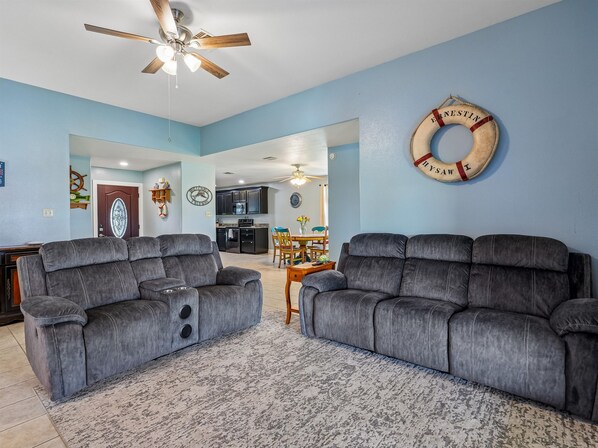 A living room with two grey sofas, a patterned rug, a ceiling fan, and a life ring decoration on the wall. - Sorry Charlie's | Waterfront Serenity (Rockport)