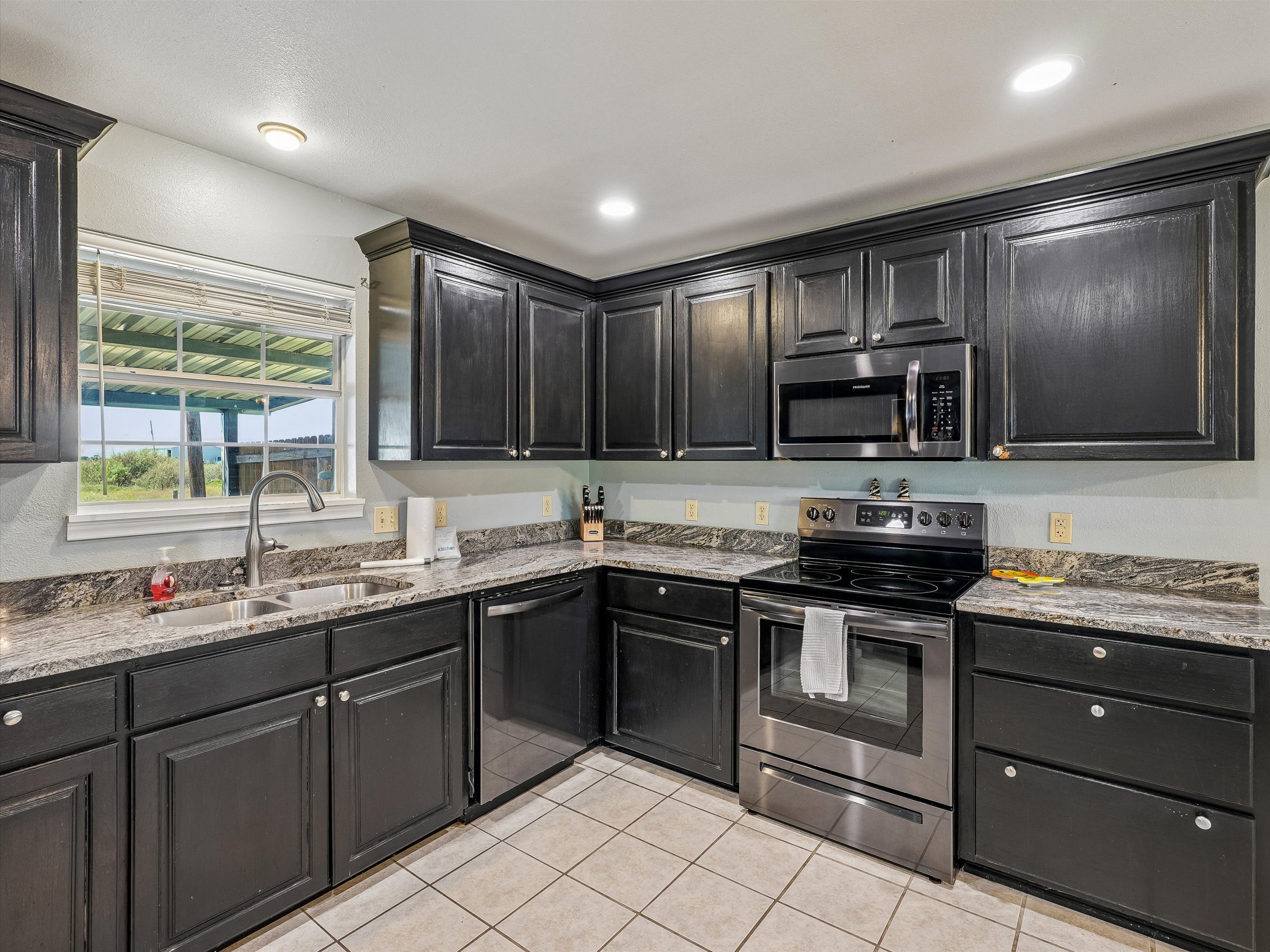 A kitchen with dark cabinets, a stainless steel stove, a microwave, and a window with a view.