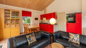 A modern kitchen with red cabinets and a built-in oven. There is a dining area with a wooden table and chairs, and a black leather sofa with floral cushions. - Holiday Home Near Beach (Oostkapelle)
