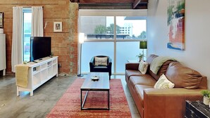A living room with a brick wall, a TV on a white stand, a brown leather sofa, a red rug, and a balcony with a view of buildings. - Tremont Meilleur 104 | A Downtown Escape (Galveston)
