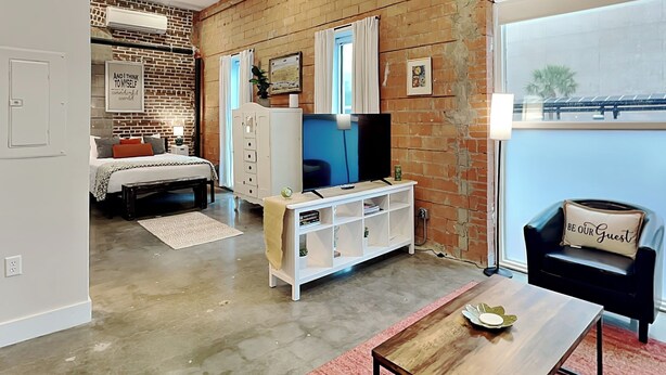 A modern living room with a brick wall, a large TV, a sofa, and a dining table. - Tremont Meilleur 104 | A Downtown Escape (Galveston)