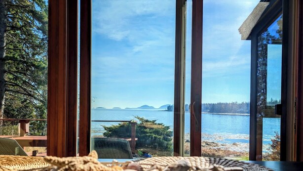 Een kamer met een groot raam met uitzicht op een meer, houten meubels en een geweven mat op de tafel.