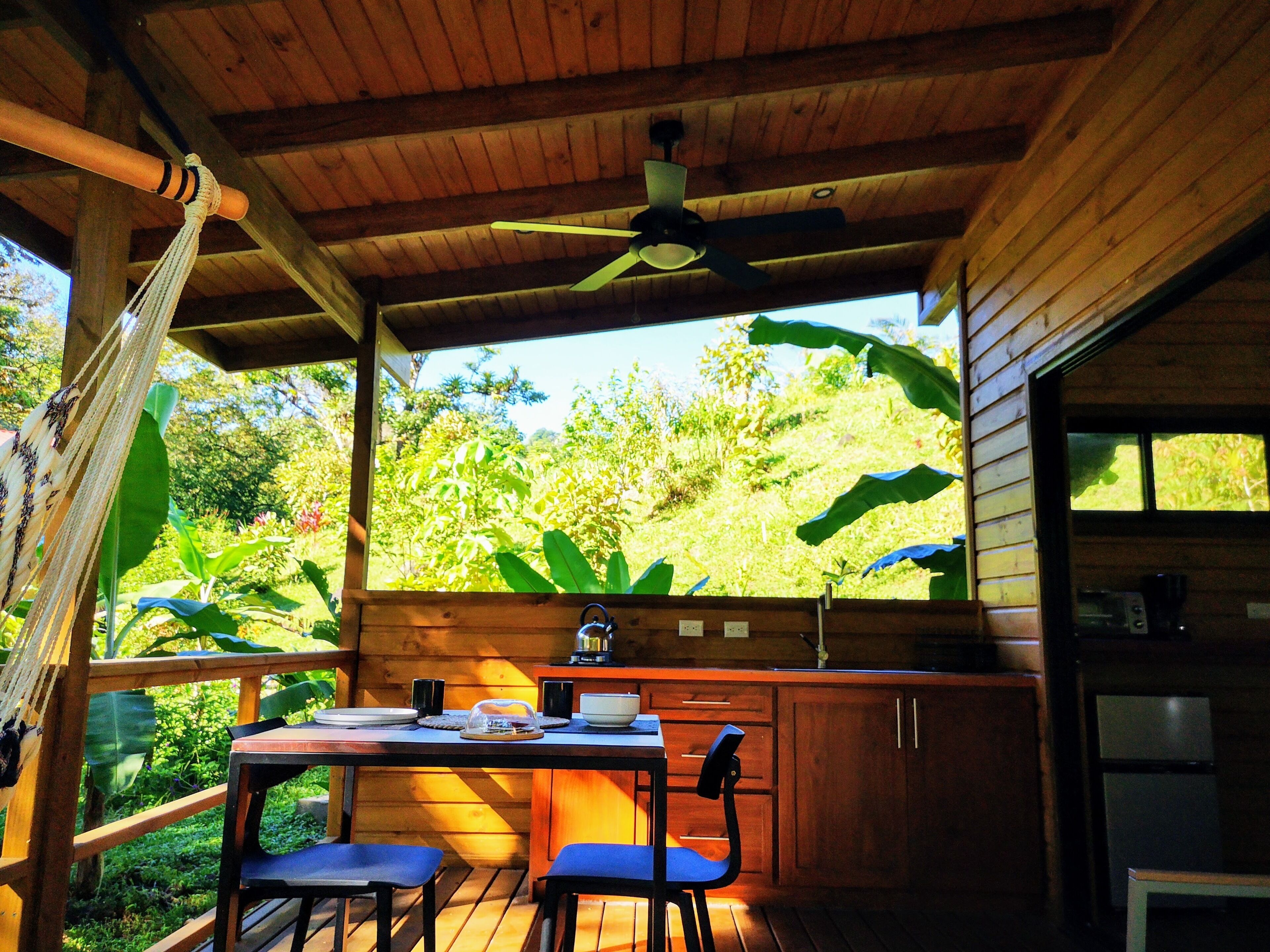 A wooden deck with a table and chairs, a ceiling fan, and a view of lush greenery.