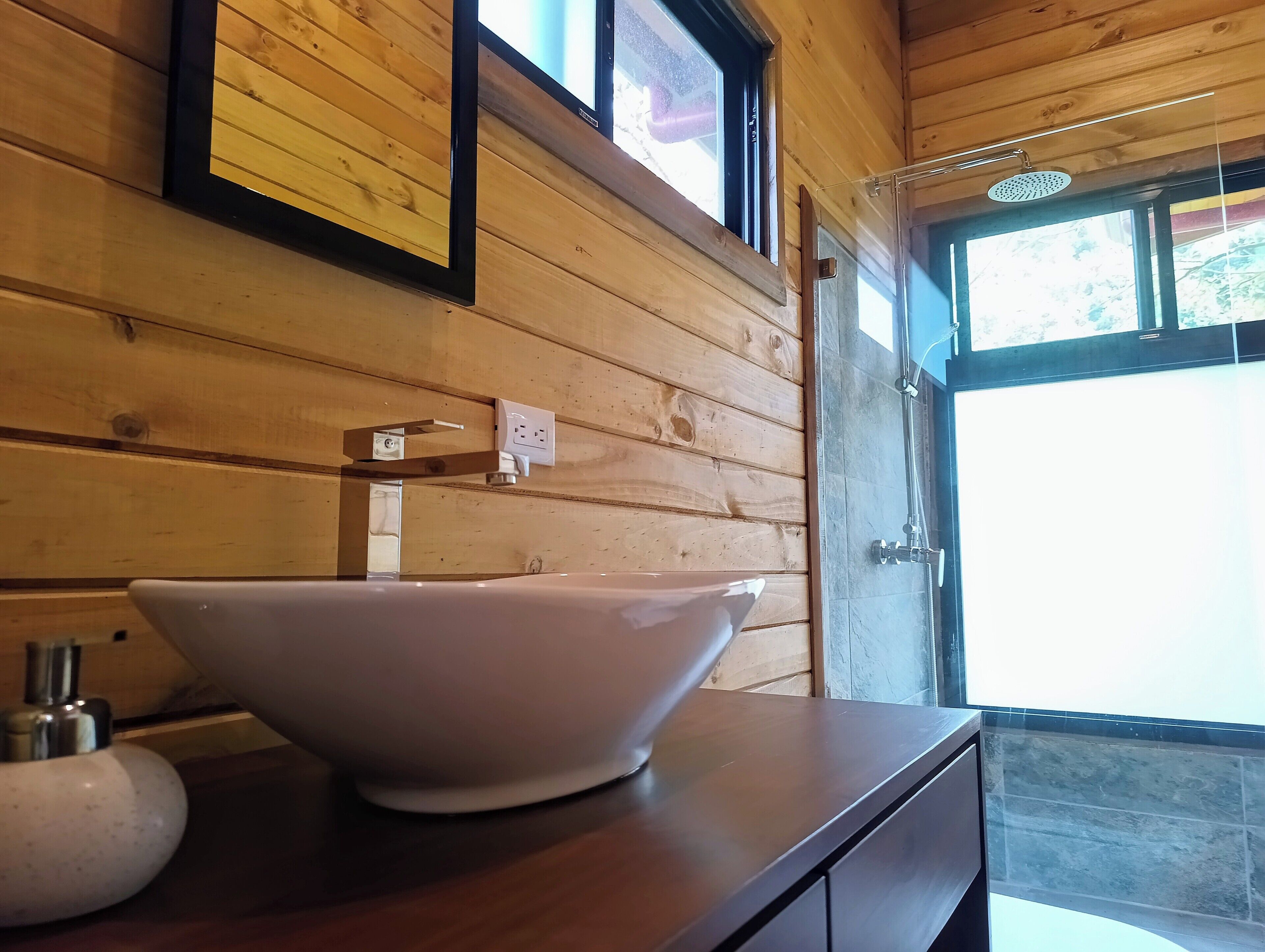 A wooden cabin interior with a large white basin on a wooden countertop, a window with a view, and a shower area.