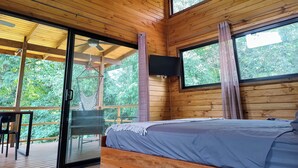 Een houten kamer met een bed, plafondventilator en grote ramen met uitzicht op groen.