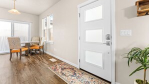 Een kamer met een eettafel, stoelen, een witte deur, een kleine plant en een kamerkleed met een patroon.