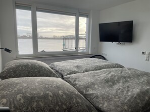 Ein Schlafzimmer mit Bett, Fernseher, Fenster mit Blick auf ein Gewässer und einem Sessel.