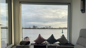 Ein modernes Zimmer mit großem Fenster und Blick auf ein Gewässer, einer Bank mit Kissen sowie einem grauen Sessel.