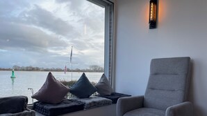 Ein modernes Zimmer mit großem Fenster und Blick auf ein Gewässer, ein grauer Sessel und eine Sitzbank mit Kissen.