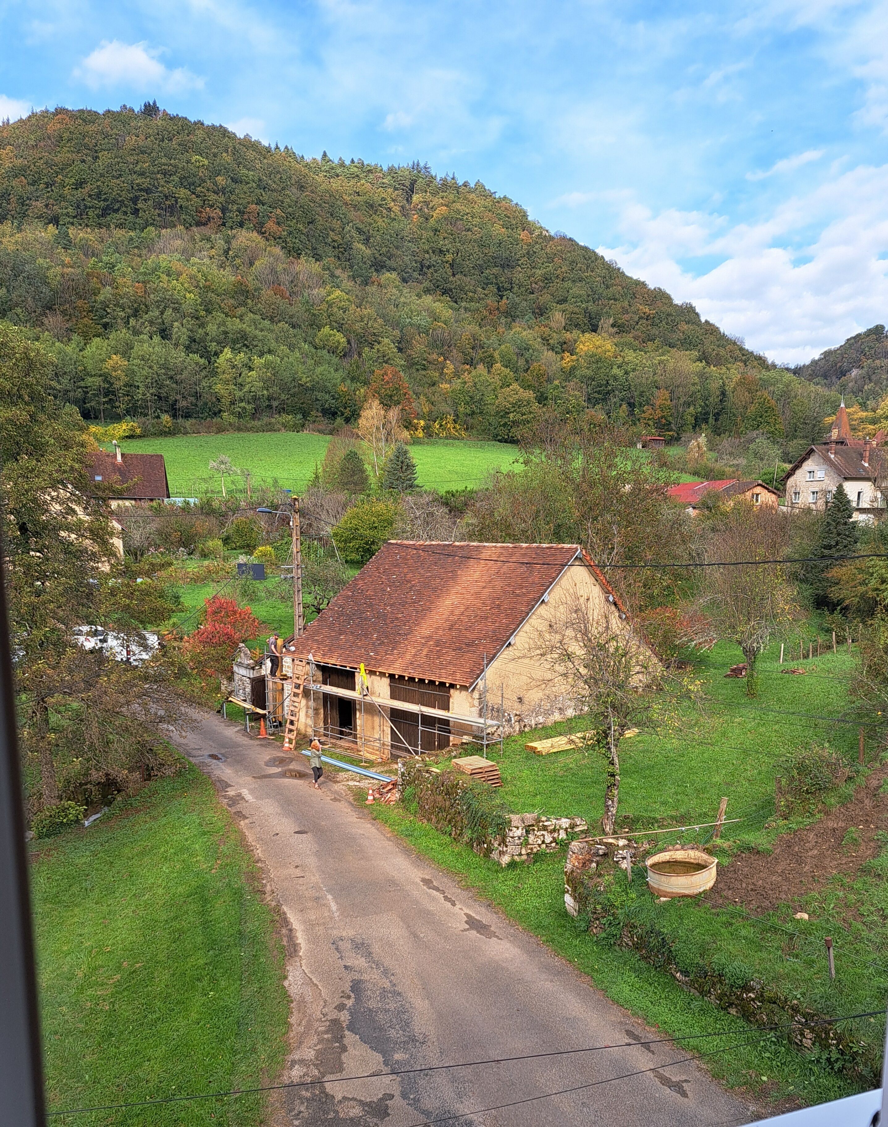 A rural landscape with a house surrounded by greenery and a hill in the background.