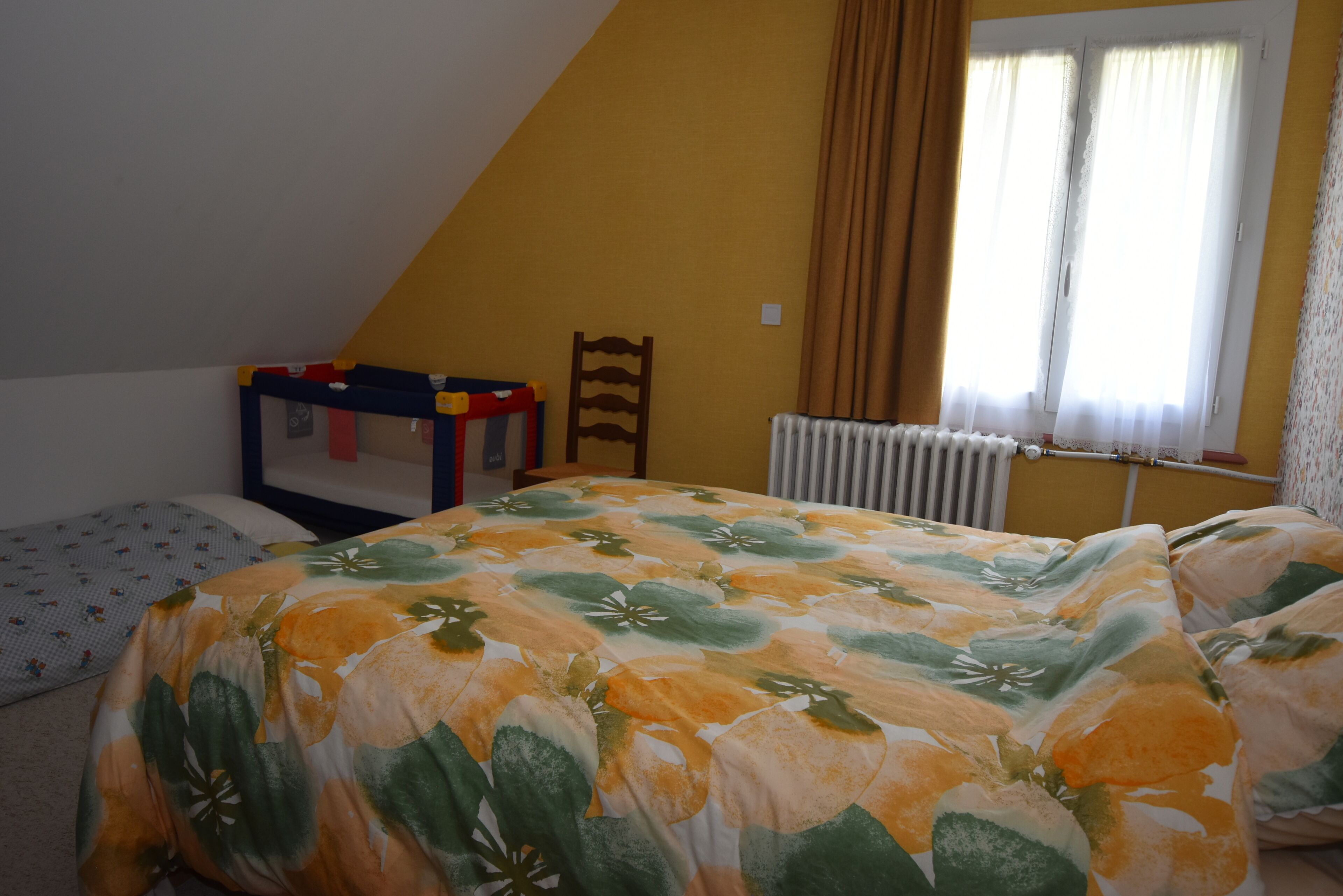 A bedroom with a sloped ceiling, a bed with a floral bedspread, a small chair, a radiator, and a window with curtains.